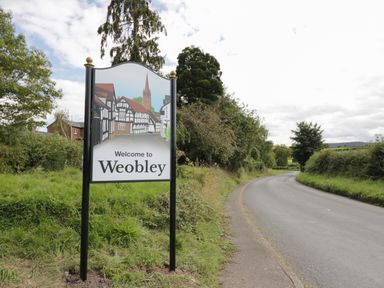 A road with a welcome to Weobley sign next to a grassy area and trees at The Folly in Weobley