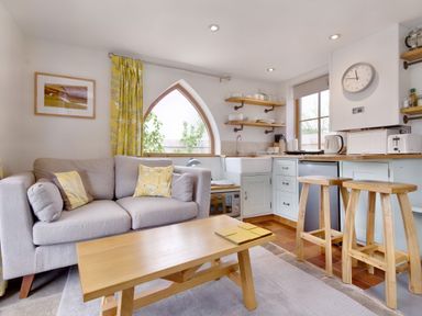 A living room with a gray sofa yellow pillows wooden coffee table and a kitchen with wooden stools and white cabinets at The Folly in Weobley