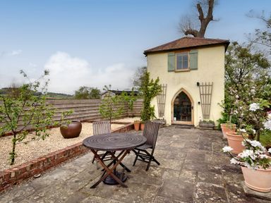 A small yellow house with green shutters and arched door with wooden table and chairs on stone patio surrounded by plants and wooden fence at The Folly in Weobley