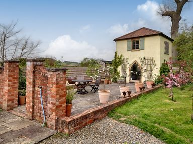 An outdoor patio area with potted plants a round table and chairs next to a small yellow two-story building at The Folly in Weobley