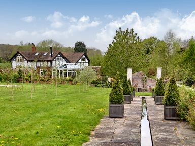 A garden pathway with potted plants leading to a brick fountain and a house in the background at The Folly in Weobley