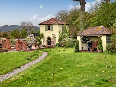 A garden with a small two-story building and a covered seating area surrounded by grass and plants at The Folly in Weobley