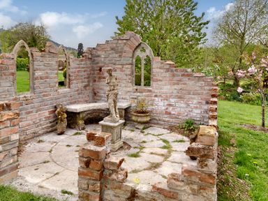 A partially ruined brick garden structure with stone benches and statues surrounded by grass and trees at The Folly in Weobley