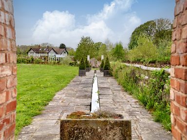 A stone pathway with a narrow water channel and potted plants leading to a house with a conservatory and surrounding lawn at The Folly in Weobley