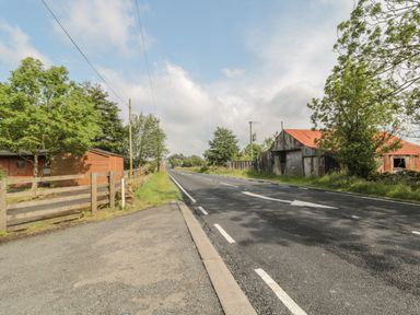 A road with a wooden fence, buildings, and trees along the sides at Vallum Kiosk in Haltwhistle