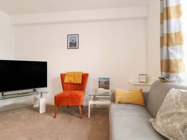 A living room with a grey sofa and orange chair with a glass TV stand and framed picture on the wall at Holly Cottage in Keswick