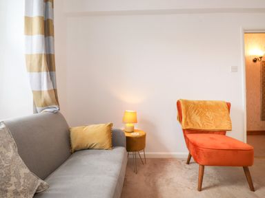 A living room with a gray sofa with cushions a small round side table with a lamp and an orange armchair with a blanket at Holly Cottage in Keswick