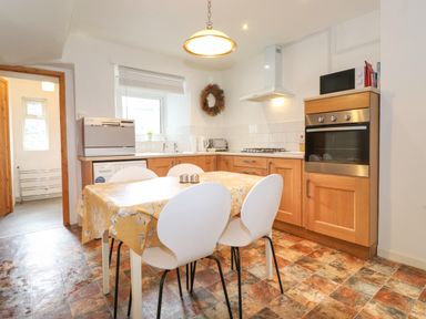 A kitchen with a table covered by a yellow tablecloth surrounded by white chairs and wooden cabinets with appliances at Holly Cottage in Keswick
