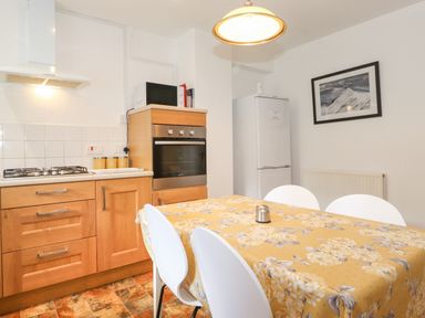 A kitchen with wooden cabinets a gas stove an oven a microwave and a refrigerator with a dining table covered by a floral tablecloth and white chairs at Holly Cottage in Keswick
