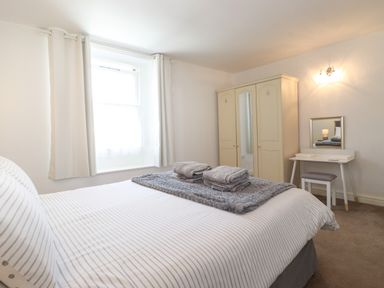 A bedroom with a bed with striped bedding folded towels on a blanket a window with curtains a wardrobe and a dressing table with a stool at Holly Cottage in Keswick