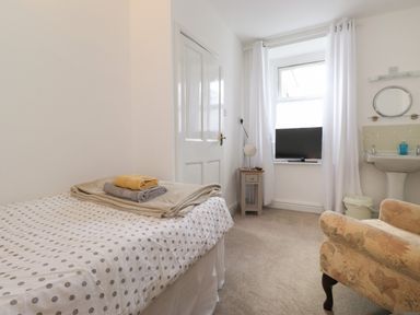 A small bedroom with a single bed towels on the bed a window with white curtains a small side table with a mirror a television a wash basin and an armchair at Holly Cottage in Keswick