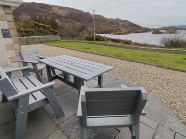 An outdoor patio with a gray table and chairs overlooking a grassy area and a body of water with hills in the background at Ranny Roe in Portsalon County Donegal