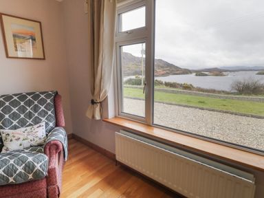 A corner of a room with an armchair covered with a quilt and cushion next to a window with beige curtains overlooking a lake and hills at Ranny Roe in Portsalon County Donegal
