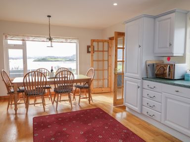 A dining area with a wooden table and chairs next to a large window and kitchen cabinets at Ranny Roe in Portsalon County Donegal
