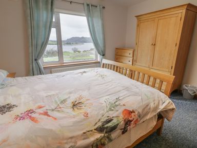 A bedroom with a wooden bed a large window with curtains a wooden wardrobe a wooden dresser and a view of a lake at Ranny Roe in Portsalon County Donegal