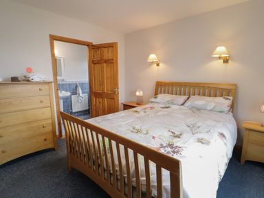 A bedroom with a wooden bed, two wall lamps, two bedside tables, a chest of drawers, and an open door showing a bathroom at Ranny Roe in Portsalon County Donegal