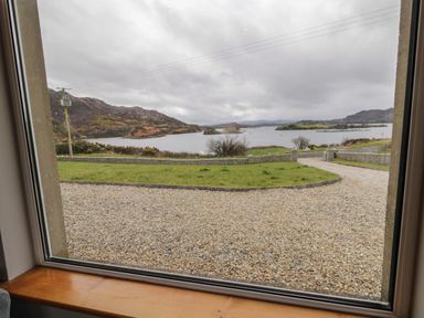 A view of a gravel driveway stone wall grassy area and a lake with hills in the background seen through a window at Ranny Roe in Portsalon County Donegal