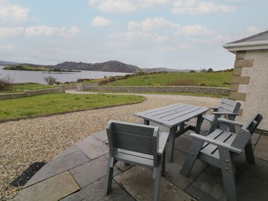 An outdoor patio with a gray table and four chairs overlooking a gravel driveway grassy area and a body of water with hills in the background at Ranny Roe in Portsalon County Donegal