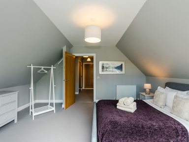 A bedroom with a bed with folded towels on a purple blanket a bedside table with a lamp a white chest of drawers a clothes rack and an open wooden door at Otter Lodge in Feshiebridge near Aviemore