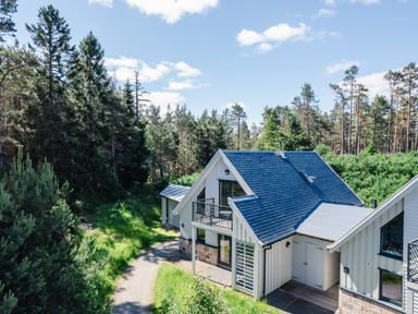 A house with a dark roof and light walls surrounded by trees and a forest path at Otter Lodge in Feshiebridge near Aviemore