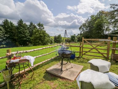 An outdoor area with a barbecue grill a wooden table with bottles and napkins a wooden fence and two stone stools with white cloths at High Rigg Shepherd's Retreat in Brampton Cumbria
