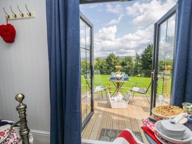 A view from inside a room looking out through open glass doors onto a wooden deck with a small table set for dining and two chairs overlooking a grassy garden with trees at High Rigg Shepherd's Retreat in Brampton Cumbria