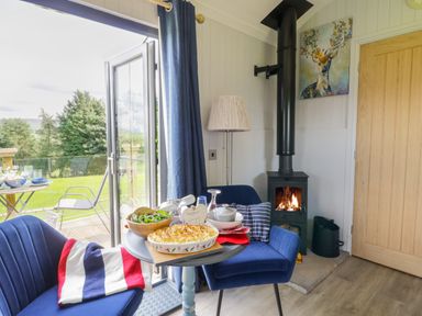 A small dining area with two blue chairs a round table with food near a wood burning stove and open door to a garden at High Rigg Shepherd's Retreat in Brampton Cumbria