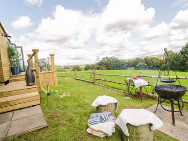 An outdoor grassy area with wooden steps leading to a deck a fire pit two stools covered with white cloth a small table with drinks and a basket with towels at High Rigg Shepherd's Retreat in Brampton Cumbria