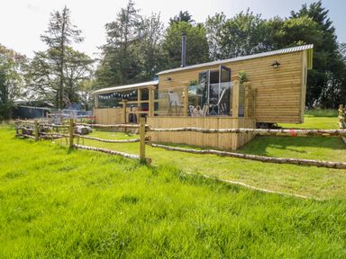 A wooden cabin with a small deck and outdoor chairs surrounded by a rustic wooden fence and green grass at High Rigg Shepherd's Retreat in Brampton Cumbria