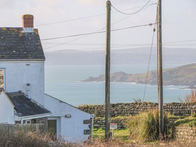 A white house near a stone wall with two wooden utility poles and a coastline in the background at Kernyk Rinsey near Porthleven