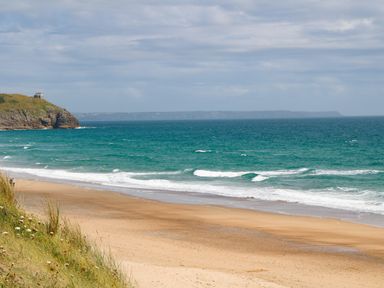 A sandy beach with ocean waves and a grassy cliff with a house at Kernyk Rinsey near Porthleven