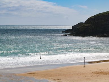 A beach with a person standing in the water and a flag on the sand near rocky cliffs at Kernyk in Rinsey near Porthleven