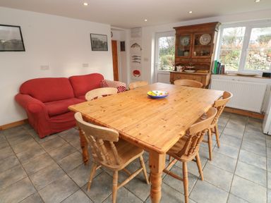 A dining area with a wooden table and six chairs next to a red sofa and wooden cabinet at Kernyk Rinsey near Porthleven