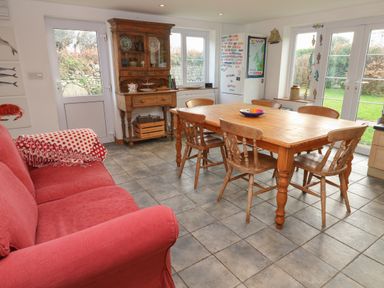 A kitchen dining area with a wooden table and chairs a red sofa a wooden cabinet and a refrigerator with windows and doors showing a garden at Kernyk in Rinsey near Porthleven
