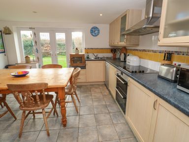 A kitchen with a wooden dining table and chairs a counter with kitchen appliances and cabinets and glass doors leading to a garden at Kernyk Rinsey near Porthleven