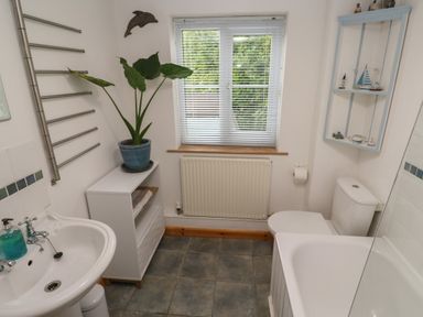A bathroom with a sink, bathtub, toilet, plant on cabinet, window with blinds and wall-mounted shelf at Kernyk in Rinsey near Porthleven