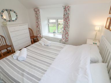 A bedroom with a bed with striped bedding and towels on it a white chest of drawers two wooden chairs a window with floral curtains and a bedside table with a lamp at Kernyk Rinsey near Porthleven