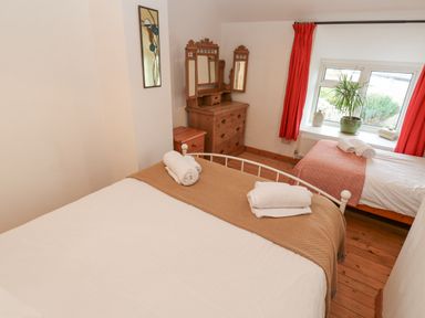 A bedroom with two beds a wooden dresser with mirror a window with red curtains and a plant on the sill at Kernyk Rinsey near Porthleven