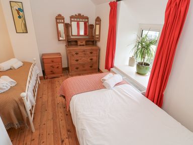 A bedroom with two single beds a wooden chest of drawers a small cabinet red curtains and a window with a plant at Kernyk in Rinsey near Porthleven