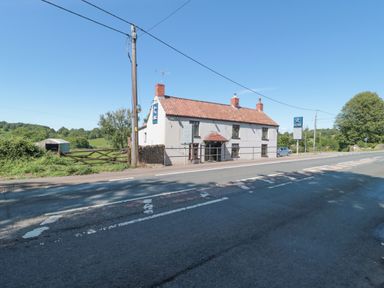 A road with a white building with a red tiled roof and signs beside green fields at The Cider Shed in Winscombe