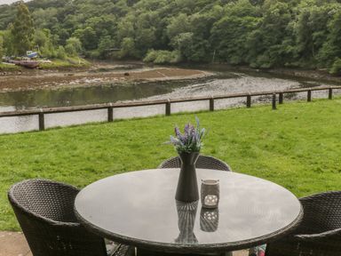 An outdoor seating area with a round glass table and wicker chairs overlooking a river and trees at Maplebeck in Whitby