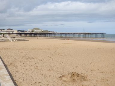 A sandy beach with a pier extending over the sea and some people walking near the water at Puffin View in Rhos-On-Sea