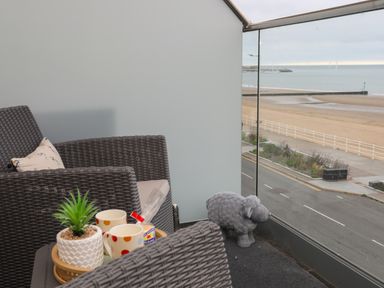 A balcony with two woven chairs a tray with cups a plant and a sheep figurine overlooking a beach at Puffin View in Rhos On Sea