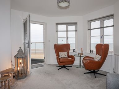 A seating area with two brown armchairs and a round glass table near windows overlooking a beach at Puffin View in Rhos-On-Sea