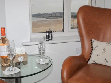 A glass table with a bottle of wine and two wine glasses next to a brown leather chair with a pillow by a window overlooking a beach at Puffin View in Rhos-On-Sea
