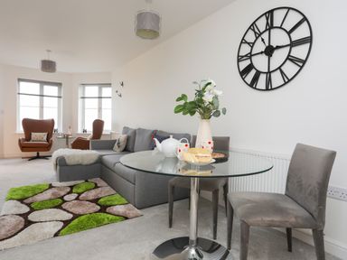 A living room with grey sofa green and brown rug glass dining table with chairs and large wall clock at Puffin View in Rhos-On-Sea