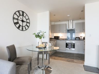 A kitchen and dining area with a glass table set with cups and snacks and a large wall clock at Puffin View in Rhos-On-Sea
