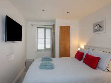 A bedroom with a bed with red pillows blue towels a wall mounted TV a window and a wooden door at Puffin View in Rhos-On-Sea