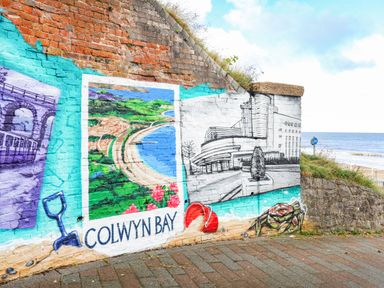 A brick wall with colorful murals including a beach scene labeled Colwyn Bay a beach bucket and crab with the sea in the background at Puffin View in Rhos-On-Sea