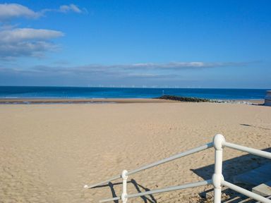 A sandy beach with a railing in the foreground and wind turbines in the sea at Puffin View in Rhos-On-Sea